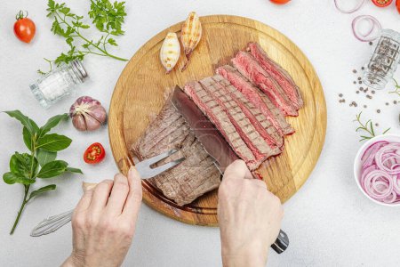 Photo for Woman cuts grilled steak. Female hands with knife and two pronged fork. Fresh herbs, spices, vegetables. Wooden cooking board, picnic or barbecue concept. Light stone concrete background, top view - Royalty Free Image