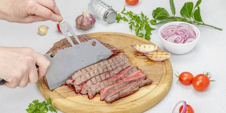 Photo for Woman cuts grilled steak. Female hands with knife and two pronged fork. Fresh herbs, spices, vegetables. Wooden cooking board, picnic or barbecue concept. Light stone background, banner format - Royalty Free Image