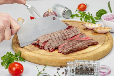Photo for Woman cuts grilled steak. Female hands with knife and two pronged fork. Fresh herbs, spices, vegetables. Wooden cooking board, picnic or barbecue concept. Light stone concrete background, close up - Royalty Free Image