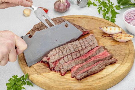 Photo for Woman cuts grilled steak. Female hands with knife and two pronged fork. Fresh herbs, spices, vegetables. Wooden cooking board, picnic or barbecue concept. Light stone concrete background, close up - Royalty Free Image