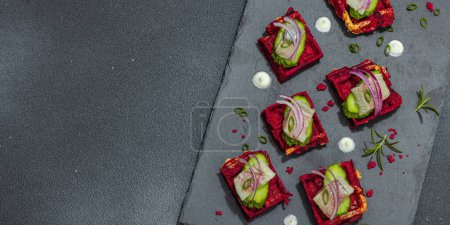 A delicious festive appetizer with herring on pieces of beet waffles with spices and sauce. Trendy hard light, dark shadow, black stone concrete background, banner format
