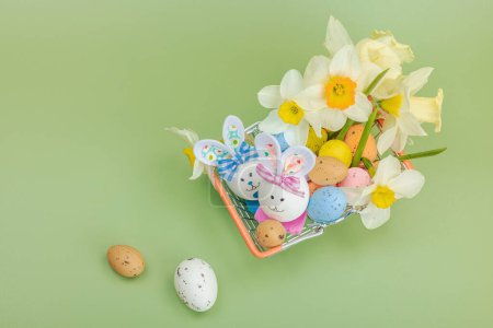 Easter sale concept. Shopping basket with festive symbols - rabbit, eggs, bird, and traditional decor. Pastel design, green background, top view