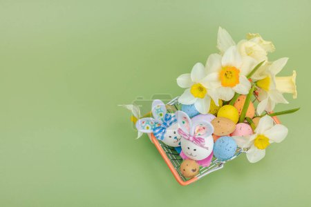 Easter sale concept. Shopping basket with festive symbols - rabbit, eggs, bird, and traditional decor. Pastel design, green background, top view