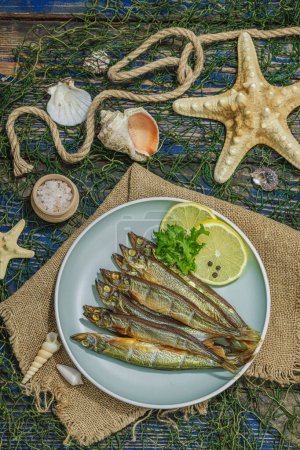Photo for Smoked smelt with fresh lemon and herbs. Salted fish with marine decor. Trendy dish, sea rope. Blue nautical wooden background, top view - Royalty Free Image