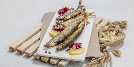 Photo for Smoked smelt with fresh lemon and herbs. Salted fish with marine decor. Trendy pallet, sea rope. Hard light, dark shadow, light stone concrete background, banner format - Royalty Free Image