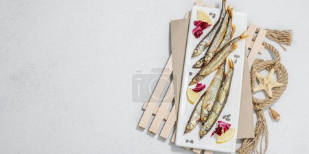 Photo for Smoked smelt with fresh lemon and herbs. Salted fish with marine decor. Trendy pallet, sea rope. Hard light, dark shadow, light stone concrete background, banner format - Royalty Free Image
