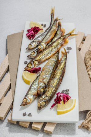 Photo for Smoked smelt with fresh lemon and herbs. Salted fish with marine decor. Trendy pallet, sea rope. Hard light, dark shadow, light stone concrete background, close up - Royalty Free Image