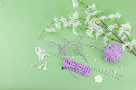 Spring knitting concept. Pattern example, traditional tools, ball of yarn, blooming cherry branch. Creative handmade flat lay, pastel green background, top view