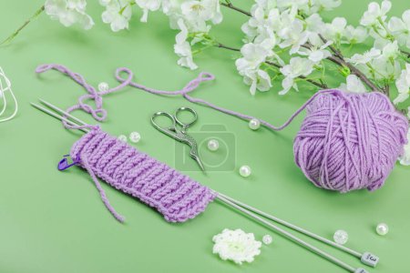 Spring knitting concept. Pattern example, traditional tools, ball of yarn, blooming cherry branch. Creative handmade flat lay, pastel green background, close up