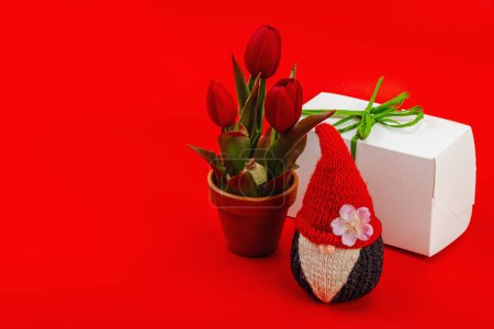 Minimalist spring greeting card with knitted gnome, tulips and gift box. Traditional festive symbol, handmade concept. Passion red background, copy space