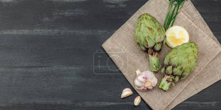 Fresh green artichokes cooking on wooden background. Traditional seasonal ingredients for healthy vegan food, flat lay. Spices, greens, dark theme, banner format