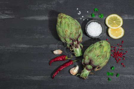 Fresh green artichokes cooking on wooden background. Traditional seasonal ingredients for healthy vegan food, flat lay. Spices, greens, dark theme, top view