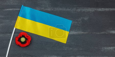 Memory Day concept. Ukrainian flag and handmade crochet poppy flower. Traditional symbols, flat lay, black wooden background, banner format