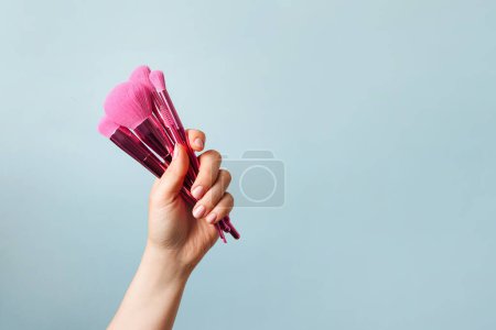 Photo for Hand holding a set of pink makeup brushes on blue background for cosmetic advertising, copy space - Royalty Free Image