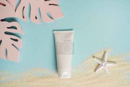 Cosmetic cream white tube mockup on blue background with sand, starfish and pink monstera leaves. Sunscreen with UVA and UVB protection