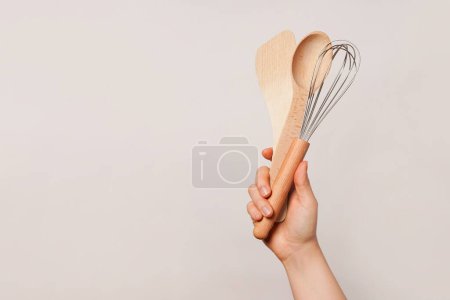 Hand holding a wooden spoon, a wooden spatula, and a whisk against a plain beige background, copy space