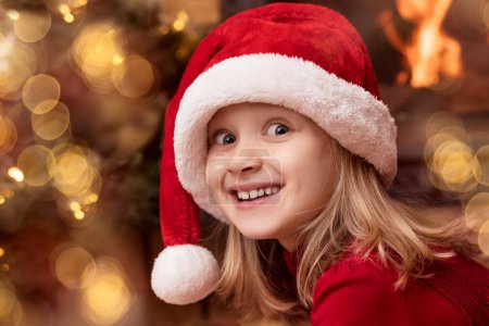 Christmas Child Girl Portrait in Santa Hat on Fireplace Background with Christmas Lights. Happy Christmas Children Holidays concept