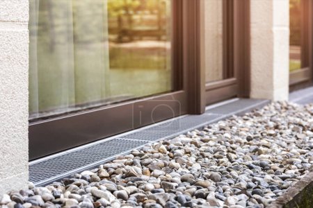 French Drain covered with Drainage Rocks and Grid. Drainage floor System for Storm water by Panoramic Window Wall of House.