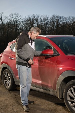 Téléchargez les photos : Homme plus âgé prendre soin annd nettoyage de voiture en plein air sur la nature parking - en image libre de droit
