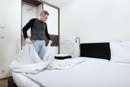 Téléchargez les photos : Homme plus âgé travaillant dans la chambre à coucher sur faire le lit - en image libre de droit