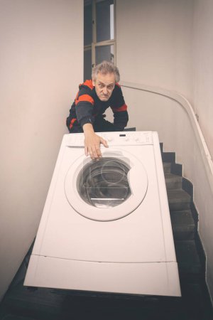 Photo for Older man in overall moving wash machine on cart in building - Royalty Free Image