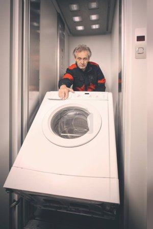 Photo for Older man in overall moving wash machine on cart with elevator - Royalty Free Image