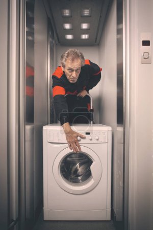 Photo for Older man in overall moving wash machine on cart with elevator - Royalty Free Image