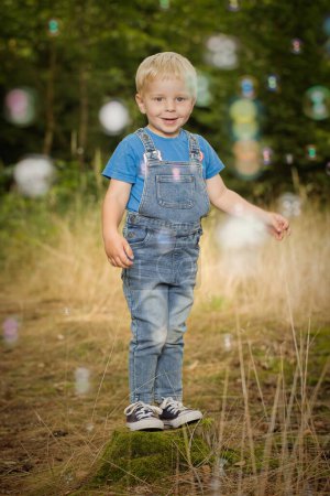 Foto de Niño pequeño vestido con jeans posando sobre muñón y sendero en el bosque - Imagen libre de derechos