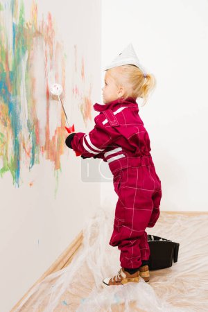 Foto de Niña rubia en overol reparando y pintando pared blanca - Imagen libre de derechos