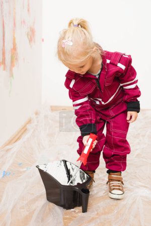 Photo for Little blonde girl in overalls repairing and painting white wall - Royalty Free Image