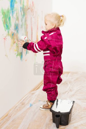 Photo for Little blonde girl in overalls repairing and painting white wall - Royalty Free Image