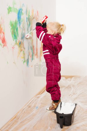 Téléchargez les photos : Petite fille blonde en salopette réparer et peindre mur blanc - en image libre de droit