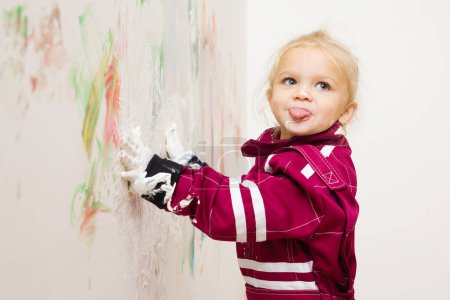 Foto de Niña rubia en overol jugando con el color blanco en la pared - Imagen libre de derechos