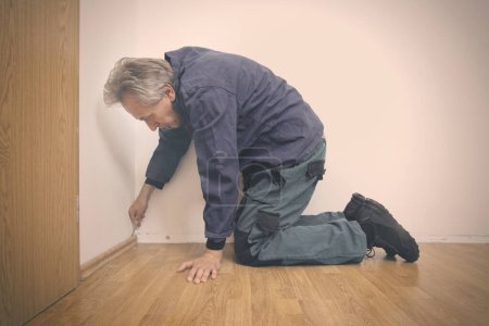 Téléchargez les photos : Homme plus âgé nettoyage et réparation du sol dans un appartement vide - en image libre de droit
