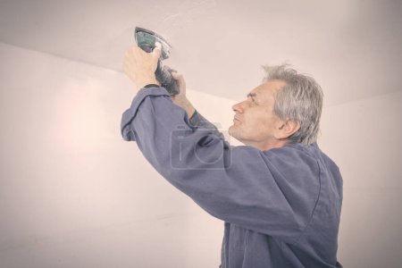 Téléchargez les photos : Homme plus âgé réparer et poncer le plafond dans un appartement vide - en image libre de droit