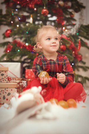 Photo for Girl in red at time of christmas day unpacking gifts near tree - Royalty Free Image