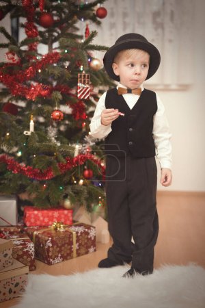 Téléchargez les photos : Garçon en chapeau melon au moment du jour de Noël déballer des cadeaux près de l'arbre - en image libre de droit
