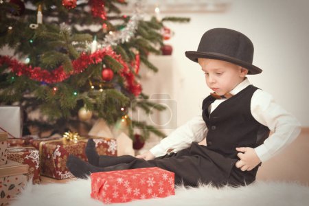 Photo for Boy in bowler hat at time of christmas day unpacking gifts near tree - Royalty Free Image