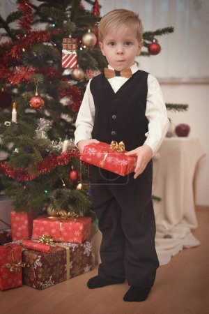 Téléchargez les photos : Garçon en robe formelle au moment du jour de Noël déballer des cadeaux près de l'arbre - en image libre de droit
