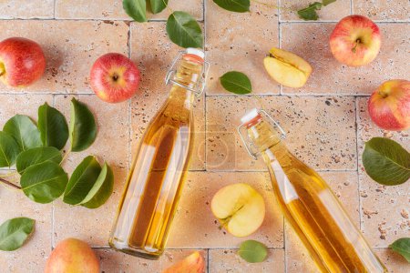 Apfelessig aus fermentierten Früchten in zwei Glasflaschen mit frischen reifen roten Bio-Äpfeln auf beigem Betongrund. Vitamin-Superfood-Drink, Gesunder Lebensstil, Draufsicht, flache Lage