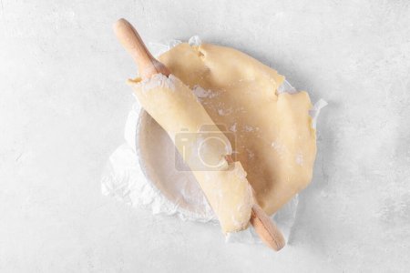 Raw crust pie dough into a baking dish with a rolling pin on a light gray background. Top view of homemade pie crust on the table. Home baking concept, pie crust recipe, hobby home bakery, top view