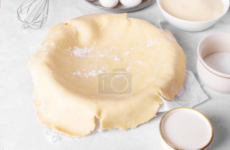 Photo for Raw crust pie dough into a baking dish with ingredient and rolling pin on a light gray background. Top view of homemade pie crust on the table. Home baking concept, pie crust recipe, hobby home bakery - Royalty Free Image