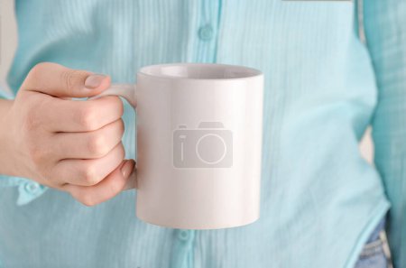 Female hand holding white mug with blank copy space for your advertising text message or promotional content. Girl in blue shirt holding white porcelain coffee or tea mug mock up