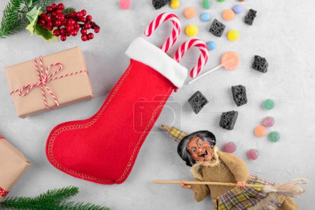 The funny witch Befana and red stocking with sweet coal and candy on light gray background. Italian Epiphany day tradition, top view, flat lay, greeting card