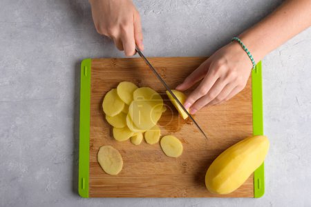 Authentic female hands cutting potatoes on wooden cutting board on kitchen table. Woman in apron cut potato, preparing food, home interior, domestic life, lifestyle, top view