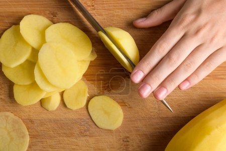 Authentic female hands cutting potatoes on wooden cutting board on kitchen table. Woman in apron cut potato, preparing food, home interior, domestic life, lifestyle, top view