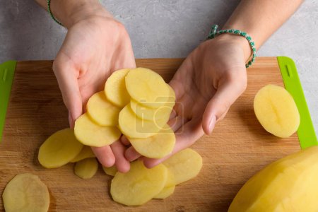 Authentic female hands is holding cutting potatoes on wooden cutting board on kitchen table. Woman in apron cut potato, preparing food, home interior, domestic life, lifestyle, top view