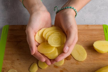 Authentic female hands is holding cutting potatoes on wooden cutting board on kitchen table. Woman in apron cut potato, preparing food, home interior, domestic life, lifestyle, top view