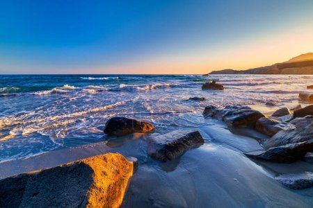 Photo for Beautiful seascape of low tidal waves, clear sunset sky, hazy hills. Clear blue and azure waters approaching washed stone buried in sand. Nature, landscape, calm and relaxing meditative view - Royalty Free Image
