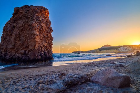 Photo for Colorful sunset over Fyriplaka beach, Milos island, Cyclades, Greece. Beautiful sky, unique lonely rock on sandy beach, low sun, sundown haze, rocky hills, sun rays, sea waves spray. - Royalty Free Image